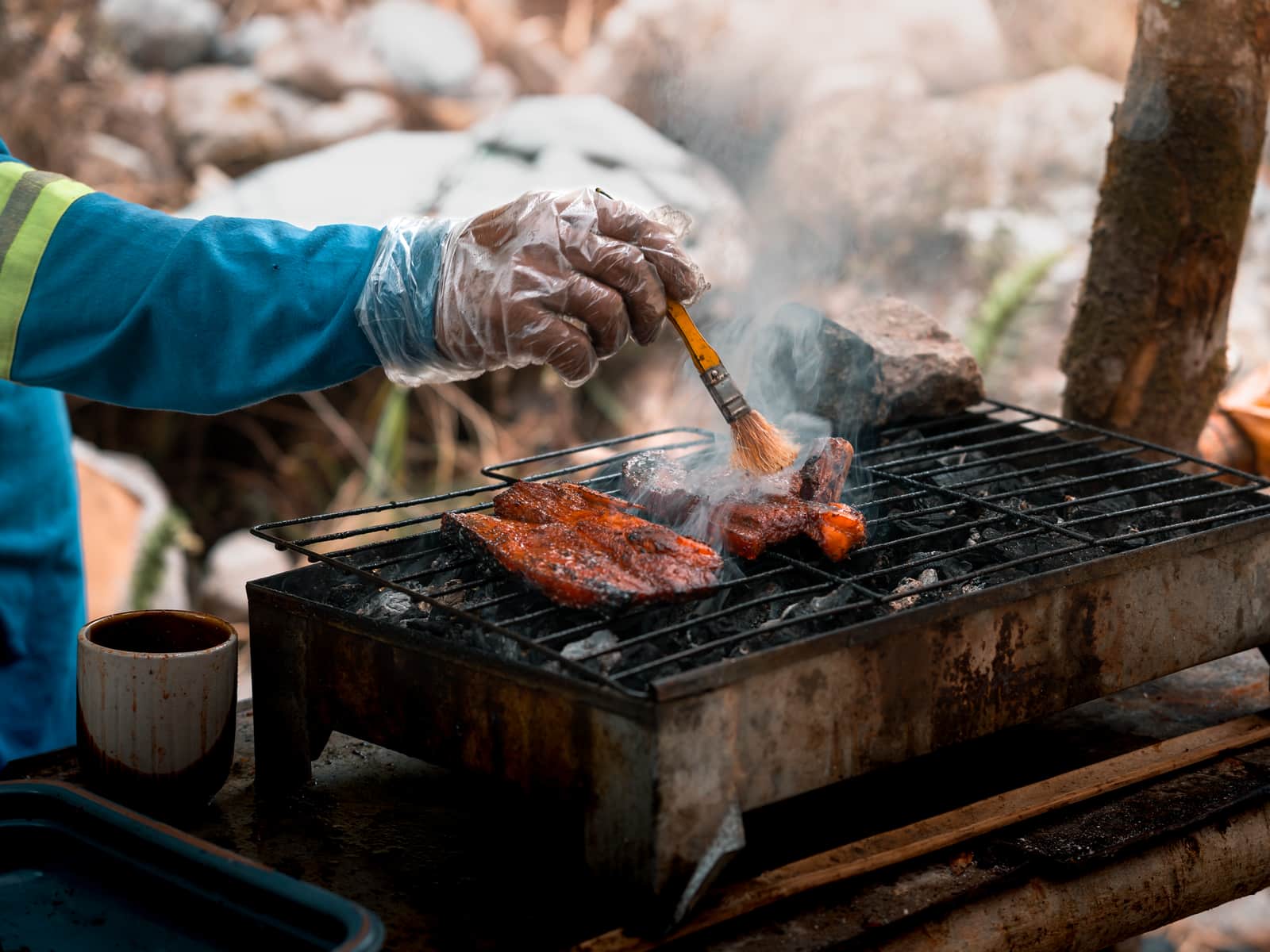 A gloved hand basting smoked meat on a charcoal grill outdoors as smoke rises