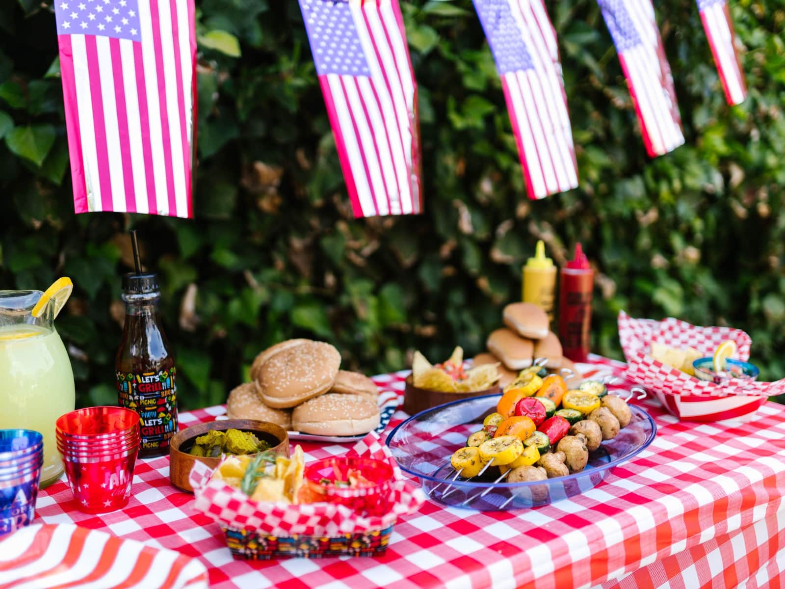 A cookout table with a checked cloth, burgers, lemonade and flag bunting